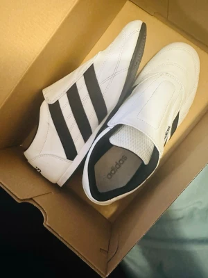 Vita Adidas slip-on sneakers - Snygga vita Adidas slip-on sneakers med svarta ränder på sidorna och svart sula. Skorna har en stilren design utan snörning och är tillverkade i läder med meshdetaljer upptill. Perfekta för en sportig och clean look. Använd två gånger bara 