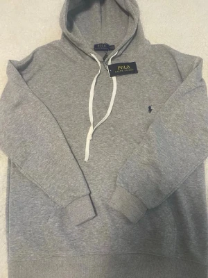 Grå hoodie från Polo Ralph Lauren - Snygg grå hoodie från Polo Ralph Lauren med klassisk huva och vita dragsnören. Broderad marinblå logga på bröstet och ribbade muddar vid ärmslut och midja. Perfekt för en chill och stilren look. Säljer den eftersom den är lite för stor.