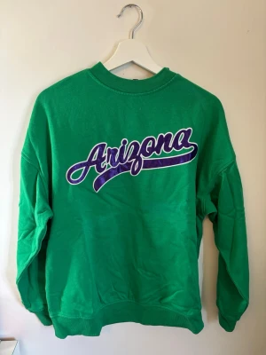 Grön sweatshirt med Arizona-tryck - Grön sweatshirt med stort Arizona-tryck i lila och vitt på bröstet. Tröjan har rund hals, ribbade muddar vid ärmslut och nederkant samt en avslappnad passform. Är väldigt stor i storleken så skulle säga att det snarare är en S. 