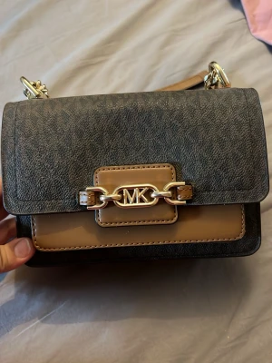 Brun och svart Michael Kors handväska - Snygg handväska från Michael Kors i konstläder med guldfärgade detaljer och kedjerem. Väskan har ett stilrent MK-emblem framtill och magnetlås. Perfekt storlek för det viktigaste och en modern accessoar till din outfit. Den är lite sliten inget som syns och lite färgad där inne av smink 