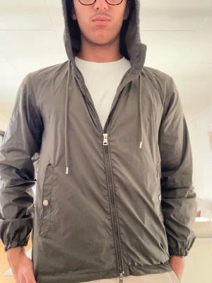Svart vindjacka med huva från Moncler - Snygg svart vindjacka från Moncler med dragkedja framtill, två fickor med knapp och justerbar huva med snören. Jackan har resår vid ärmsluten och en diskret Moncler-logga på vänster ärm. Perfekt för blåsiga dagar och enkel att matcha.