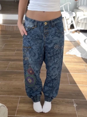 Desigual Denim baggy hippie jeans med broderier – strl 28  - Unika baggy jeans i mörkblå denim med blommigt mönster och färgglada broderier på ena benet. Resår vid benslut och flera fickor med dragkedja och detaljer. Perfekta för dig som gillar en avslappnad och kreativ stil.