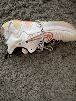 Nike vita sneakers/basketskor med färgglada detaljer - Säljer ett par vita Nike sneakers/ Gannis med vågig sula och färgglada detaljer. Ena skon har blå swoosh och den andra röd, båda med svart och vita snören och svarta detaljer vid hälen. Skorna har en modern, sportig look och är tillverkade i textil och syntet. Använda 1-3 gånger så väldigt bra skick men säljer pågrund av att de inte blir använda så någon annan kan få användning av dem. 