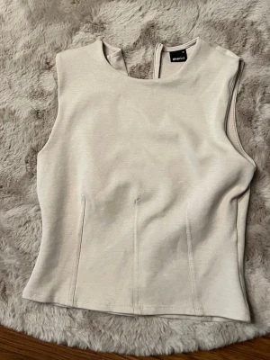 Beige ärmlös topp från Vero Moda - Säljer en stilren beige topp från Vero Moda. Toppen är ärmlös med rund halsringning och har en diskret dragkedja i nacken. Materialet känns mjukt och stretchigt, perfekt för att matcha med olika outfits.