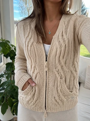 Beige stickad kofta från Odd Molly - Säljer en beige stickad kofta från Odd Molly med dragkedja framtill och snygga flätstickade detaljer. Koftan har rund halsringning, långa ärmar och ribbade muddar. Perfekt för lager på lager och ger en mysig känsla till din outfit.