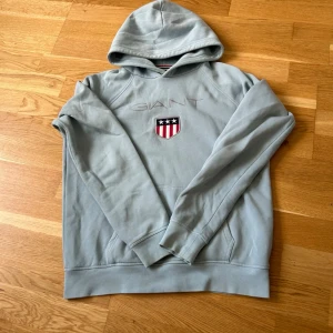 Ljusblå hoodie från GANT - Snygg ljusblå hoodie från GANT med broderad logga och ett sköldmärke med stjärnor och ränder på bröstet. Klassisk huva och ribbade muddar vid ärmslut och nederkant. Perfekt för en avslappnad och stilren look.