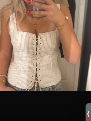 Vit randig korsettopp med snörning - Supersnygg vit korsettopp med tunna svarta ränder och snörning framtill. Toppen har breda axelband och markerad midja för en riktigt smickrande siluett. Perfekt att styla med jeans eller kjol för en trendig look.