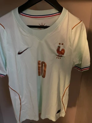 Frankrike fotbollströja VM 2026 MBAPPE - Snygg vit fotbollströja med bronsfärgade detaljer och nummer 10 på bröstet. Tröjan har Frankrikes landslagsemblem med tupp och två stjärnor, samt VM 2026-märke på ärmen. V-ringad hals och randiga muddar i blått, vitt och rött. Perfekt för fotbollsfans!