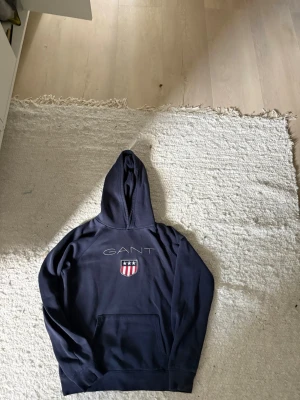 Mörkblå hoodie från GANT - Snygg mörkblå hoodie från GANT med broderad logga och amerikanskt sköldmärke på bröstet. Klassisk känguruficka framtill och huva. Perfekt för en avslappnad och sportig stil.