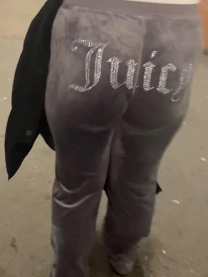 Gråa Juicy Couture mjukisbyxor - Säljer ett par gråa mjukisbyxor från Juicy Couture i mjuk velour. Byxorna har raka ben och dekoreras med glittrande stenar som stavar 'Juicy' över bakfickan. Perfekta för en chill och trendig look.
