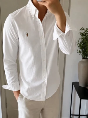 Vit skjorta från Ralph Lauren Slim Fit - Klassisk vit skjorta från Ralph Lauren i slim fit-modell. Skjortan har långa ärmar, knappar framtill och den ikoniska broderade loggan på bröstet. Tillverkad i mjuk bomull för en clean och stilren look.