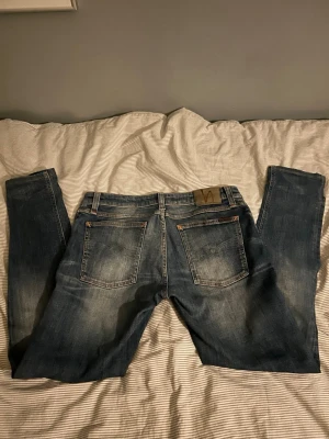 Nuddie jeans  - Säljer ett par blå slim fit jeans från Nudie Jeans med snyggt slitna detaljer och klassiska fem fickor. Jeansen har en skinnpatch med logga bak i midjan och är tillverkade i denim med en tvättad look. Perfekta för dig som gillar en avslappnad men stilren stil. Hör av er vid frågor😃