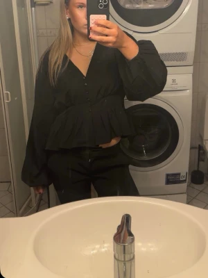 Svart volangblus med knappar - Snygg svart blus med volang nedtill och knappar framtill. Blusen har v-ringning och långa puffiga ärmar som ger en trendig look. Perfekt att styla med både jeans och kostymbyxor för en cool outfit.