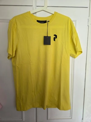 Gul t-shirt strl M  - Snygg gul t-shirt med svart geometrisk logga på bröstet och ryggen. Klassisk rund hals och korta ärmar. Tillverkad i mjuk bomull som känns skön mot huden. Perfekt för dig som vill sticka ut med en färgglad och enkel stil.
