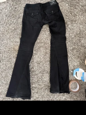 Svarta utsvängda jeans - Säljer ett par svarta jeans med utsvängda ben och klassiska femficksdetaljer. Jeansen har snygga bakfickor med lock och knapp samt låg midja. Perfekta för dig som gillar en trendig och stilren look.