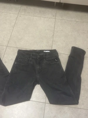 Replay Svarta/ grå slim fit jeans - Säljer ett par svarta slim fit jeans med klassisk femficksdesign och knappgylf. Jeansen har en diskret tvättad look och raka ben. Perfekta för dig som gillar en enkel och stilren stil. Jensen är knappt använda, och dom är som nya, storleken är 30-30 