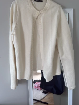 Krämvit kofta med rund hals - En stilren krämvit kofta med rund halsringning och ribbade kanter. Koftan har lång ärm och stängs med en knapp upptill. Perfekt att slänga över en topp för en enkel och clean look.