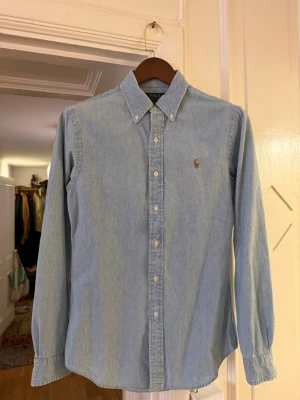 Ljusblå skjorta från Polo Ralph Lauren - Klassisk ljusblå skjorta från Polo Ralph Lauren med button-down krage och broderad logga på bröstet. Skjortan har långa ärmar, vita knappar och är tillverkad i mjuk bomull. Perfekt för en clean och stilren look.