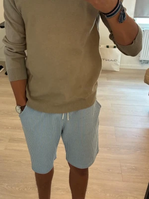 Beige stickad tröja med rund hals - Klassisk beige stickad tröja med rund hals och ribbade muddar vid ärmslut och nederkant. Tröjan har en enkel och clean look som är lätt att matcha till olika stilar. Perfekt för dig som gillar basic och tidlösa plagg.