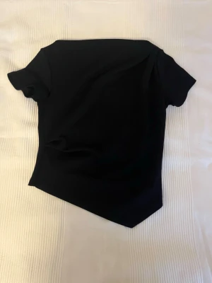 Svart asymmetrisk topp - Snygg svart topp med korta ärmar och unik asymmetrisk nederkant. Enkel och stilren design som passar till mycket. Tillverkad i mjukt material som känns skönt mot huden.
