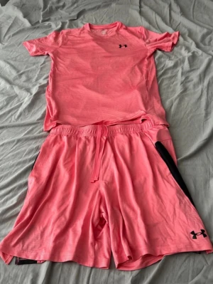 Rosa träningsset från Under Armour - Snyggt rosa träningsset från Under Armour med t-shirt och shorts. Setet är gjort i ett lätt och snabbtorkande material, perfekt för träning. T-shirten har en diskret logga på bröstet och shortsen har elastisk midja med dragsko samt svarta detaljer på sidorna. Shortsen är i storlek L och t shirten i storlek S, båda är oanvända. 