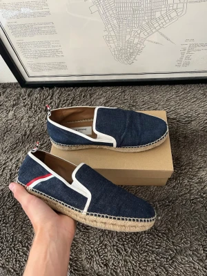 Thom Browne Mörkblå denim espadrillos - Snygga loafers i mörkblå denim med vit kant och dekorativ röd, vit och blå detalj på sidan. Skorna har en klassisk espadrillesula i flätad jute och slip-on design. Perfekta för en avslappnad och trendig look.
