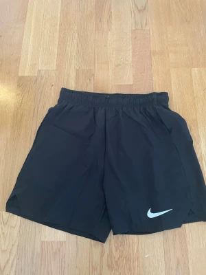 Svarta träningsshorts från Nike - Snygga svarta träningsshorts från Nike med elastisk midja och diskret Nike-logga på benet. Shortsen har en sportig look och är tillverkade i lätt och snabbtorkande syntetmaterial, perfekta för aktiva dagar.