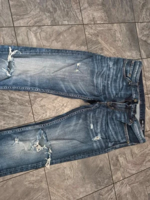 Blå slitna jeans med hål - Säljer ett par blå jeans med slitningar och hål på både fram- och baksida. Jeansen har en klassisk femficksmodell, normal midja och smal passform. Materialet är denim i bomull och de har en cool, tvättad look med tydliga slitningar.