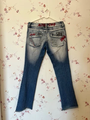 Blå jeans med nitar och brodyr - Snygga 2000s jeans washed out, dekorerade med silverfärgade nitar på bakfickorna och en röd broderad trollslända. Jeansen är utsvängda längst ner. Ett bältesband har gått upp men går att sys på igen snabbt och enkelt. Står storlek XL men skulle säga de är Storlek M på sin höjd L då de är stretchiga