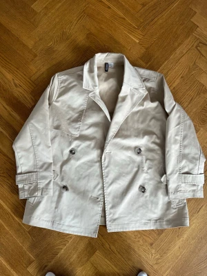 Beige trenchcoat  - Stilren beige trenchcoat med klassisk dubbelknäppning och bred krage. Jackan har justerbara remmar vid ärmsluten och en snygg stormflik på ryggen. Tillverkad i ett slätt bomullsliknande material som ger en tidlös look.