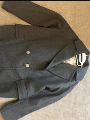 Mörkgrå ullkappa från H&M - Stilren mörkgrå kappa från H&M i storlek 38. Kappan har två knappar framtill, klassisk krage och två fickor. Materialet är ullblandning med ljus innerfoder. Perfekt för kyligare dagar och ger en clean look till alla outfits.