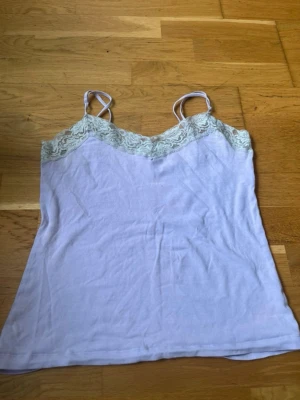 Lila  linnetopp med spetsdetaljer - Säljer en söt lila linnetopp med tunna axelband och spets upptill. Toppen har en v-ringad form framtill och spetsen ger en romantisk touch. Perfekt att styla till jeans eller kjol för en trendig look.