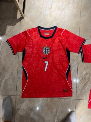 England röd fotbollströja Saka #7 - Snygg röd England fotbollströja från Nike med nummer 7 och Saka på ryggen. Tröjan har korta ärmar, svart och vit detalj längs sidorna och Englands landslagsemblem på bröstet. Perfekt för match eller träning.