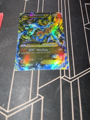 Pokémonkort Mega Charizard EX wild blaze - Holografiskt Pokémonkort med Mega Charizard EX-motiv. Kortet har en färgstark och glansig yta med blå, svart och gula detaljer samt text i guld. Perfekt för samlare eller dig som gillar Pokémon och vill ha något unikt i din kortsamling.