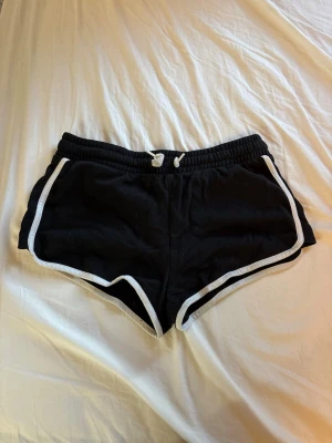 Svarta sportiga shorts med vit kant - Svarta shorts med vit kant och snörning i midjan. De har en sportig look med elastisk midja och är perfekta för träning eller chill. Materialet känns mjukt och bekvämt, och passformen är normal. Klassisk design som funkar till många stilar.