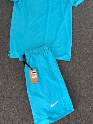  Träningsset från Nike - Snyggt turkost träningsset från Nike med t-shirt och shorts. T-shirten har korta ärmar och rund hals, medan shortsen har resår i midjan och en vit Nike-logga på benet. Perfekt för gymmet eller löprundan.
