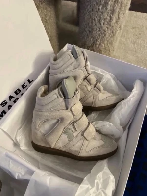 Beige höga sneakers Isabel Marant - Säljer ett par beige höga sneakers från Isabel Marant med kilklack och kardborreband. Skorna har en unik design med paneler i mocka och textil, samt en mörkare sula. Perfekta för dig som vill ha en trendig och bekväm look.