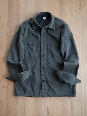 Olivgrön overshirt med fickor - Stilren olivgrön overshirt i kraftigt bomullstyg. Klassisk rak passform med två stora bröstfickor och knäppning framtill. Perfekt lager-på-lager-plagg med avslappnad krage och långa ärmar. Passar till många olika stilar.