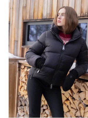 Short Black Puffer Jacket - The images show a woman wearing a short, black puffer jacket with a hood. The jacket has a front zipper closure and horizontal quilting. The first image shows the front and side view, with the woman outdoors in a winter setting, wearing black gloves and pants. The second image shows the back view of the jacket indoors, highlighting the hood and the jacket's cropped length.
