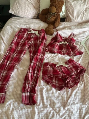 Röd- och vitrutiga pyjamas-set - Säljer ett mysigt pyjamas-set i röd- och vitrutigt mönster. Setet består av ett par långa byxor, ett linne med tunna axelband och ett par shorts med volangkant. Alla delar har en söt rosett framtill. Perfekt för chillkvällar hemma. Från Gina tricot