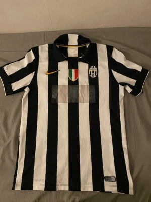 Juventus svartvit fotbollströja Nike - Juventus officiella matchtröja med klassiska svartvita ränder, gul Nike-logga och klubbmärke på bröstet. Italienska mästerskapsmärket sitter mitt på bröstet. Tröjan har krage och är tillverkad i ett lätt, andningsbart material.