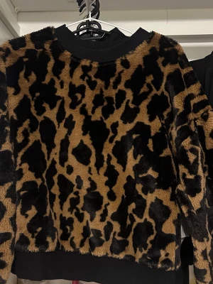 Leopardmönstrad fluffig stickad tröja - Cool stickad tröja med leopardmönster i svart och brunt. Tröjan är fluffig och har svarta muddar vid hals, ärmslut och nederkant. Perfekt för dig som vill sticka ut med en trendig och mysig look.