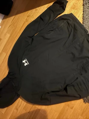 Svart hoodie från Under Armour - Svart hoodie från Under Armour med vit logga på bröstet. Klassisk modell med huva och långa ärmar. Perfekt för en sportig och avslappnad stil. Materialet känns mjukt och bekvämt mot huden.