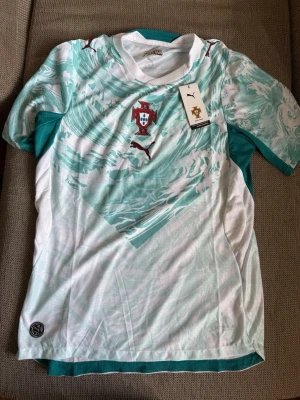 Portugal fotbollströja Puma turkos - Säljer en Portugal fotbollströja från Puma i vitt och turkost med abstrakt mönster. Tröjan har korta ärmar, rund hals och Portugal-emblemet på bröstet. Perfekt för dig som älskar fotboll och vill ha en snygg supportertröja.