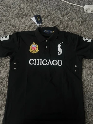 Svart pikétröja med Chicago-tryck - Svart pikétröja från Polo Ralph Lauren med vit broderad logga och texten CHICAGO på bröstet. Tröjan har krage, knappar framtill och detaljerade broderier i vitt och gult. Klassisk och stilren modell med korta ärmar. Det ser är XL med stitter som en L
