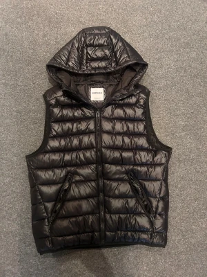 Svart dunväst med huva från Bershka - Snygg svart dunväst från Bershka med huva och dragkedja framtill. Västen har två snedställda fickor med dragkedja och quiltad design. Perfekt för lager på lager och streetstyle. Materialet är glansigt och lätt, vilket ger en modern look.