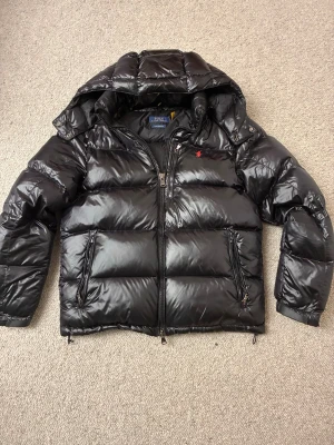 Ralph Lauren puffer i väldigt fint skick - Säljer en riktigt snygg Ralph Lauren pufferjacka i blanksvart utförande med det klassiska röda märket på bröstet. Jackan är sparsamt använd och i väldigt fint skick, perfekt vadderad för att hålla dig varm och stilren under hela vintern. Blanksvart med röd logga. Väldigt fint skick, varm och skön för vintern 