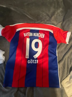 Bayern München matchtröja Götze #19 - Säljer en Bayern München matchtröja med Götze och nummer 19 på ryggen. Tröjan har röda och blå vertikala ränder, vita detaljer och klubbmärke på bröstet. Perfekt för fotbollsfans som vill visa sitt stöd för laget.