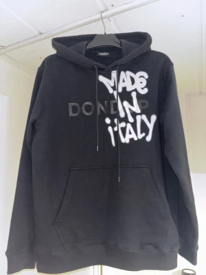 Svart Dondup hoodie med tryck - Svart hoodie från Dondup med stor vit text 'MADE IN ITALY' i graffiti-stil på bröstet och diskret Dondup-logga i svart. Klassisk känguruficka framtill, huva med dragsko och ribbade muddar. Perfekt för en streetwear-look.