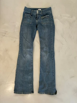 Blå bootcut jeans från Nelly - Snygga blå bootcut jeans från Nelly med dubbla knappar i midjan och två stora fickor framtill. Jeansen har markerade sömmar längs benen och är tillverkade i klassisk denim. 